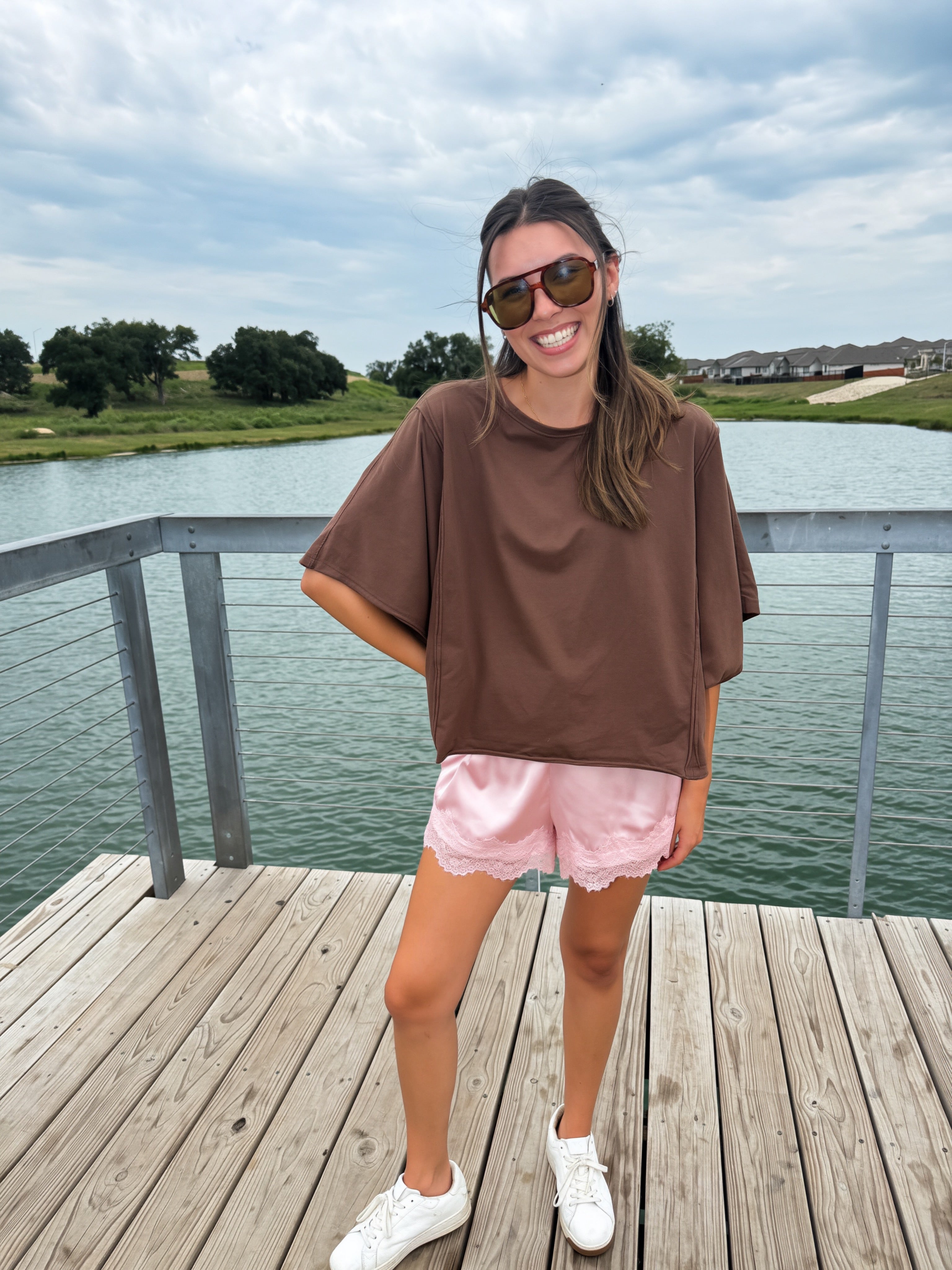 A woman stands on a wooden dock by a lake wearing an oversized brown t-shirt, light pink shorts, white sneakers, and sunglasses.