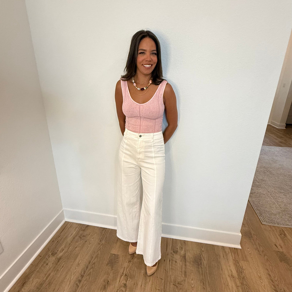 Woman wearing a pink top and white pants standing against a white wall.