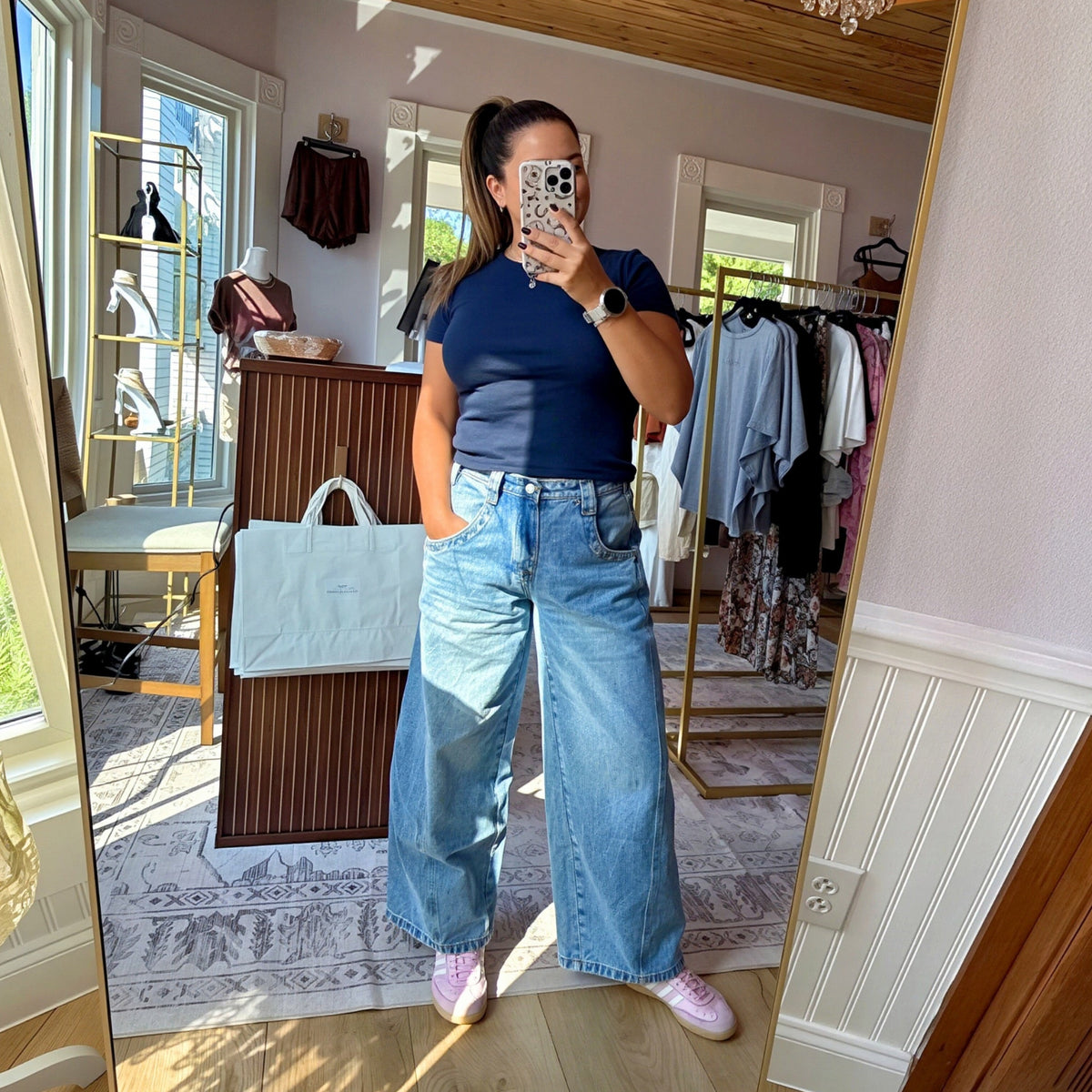 A woman takes a mirror selfie in a boutique showing a pair of wide-leg light blue denim jeans, a navy blue t-shirt, and pink sneakers. The boutique has light wood floors, a wooden ceiling with a crystal chandelier, and clothing racks in the background.