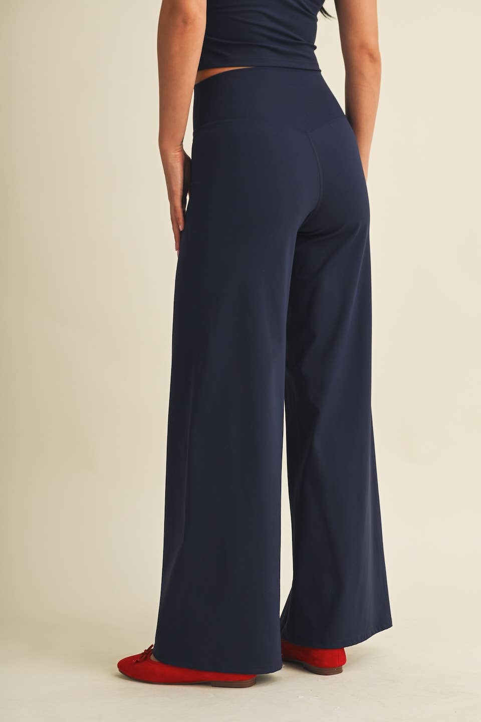 A model shown from the back wearing high-waisted, wide-leg navy blue pants and red shoes, against a plain beige studio background.