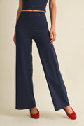 A model wearing navy blue wide-leg pants with a high waist, paired with red flats, shown from the waist down against a solid beige background.