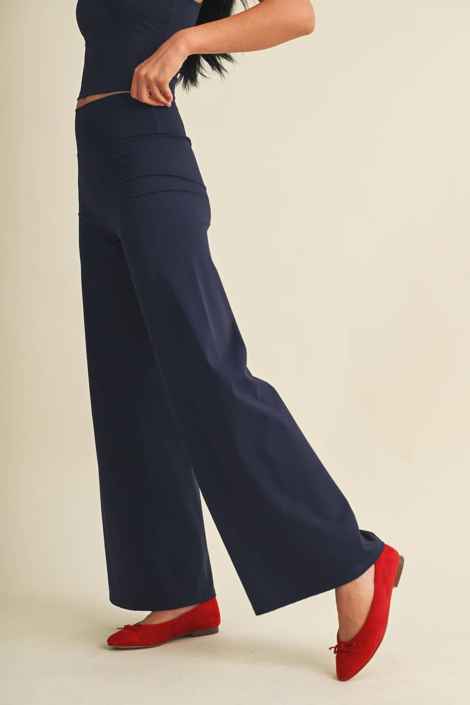 A model wearing navy blue wide-leg trousers and red pointed flats, shown from the waist down against a plain beige studio background.
