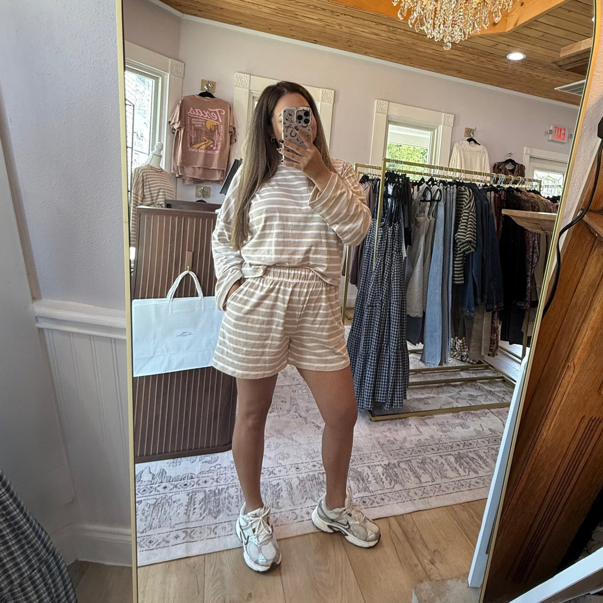 A woman takes a mirror selfie in a boutique clothing store, wearing a beige and white striped matching lounge set consisting of a long-sleeve top and shorts, paired with white sneakers.
