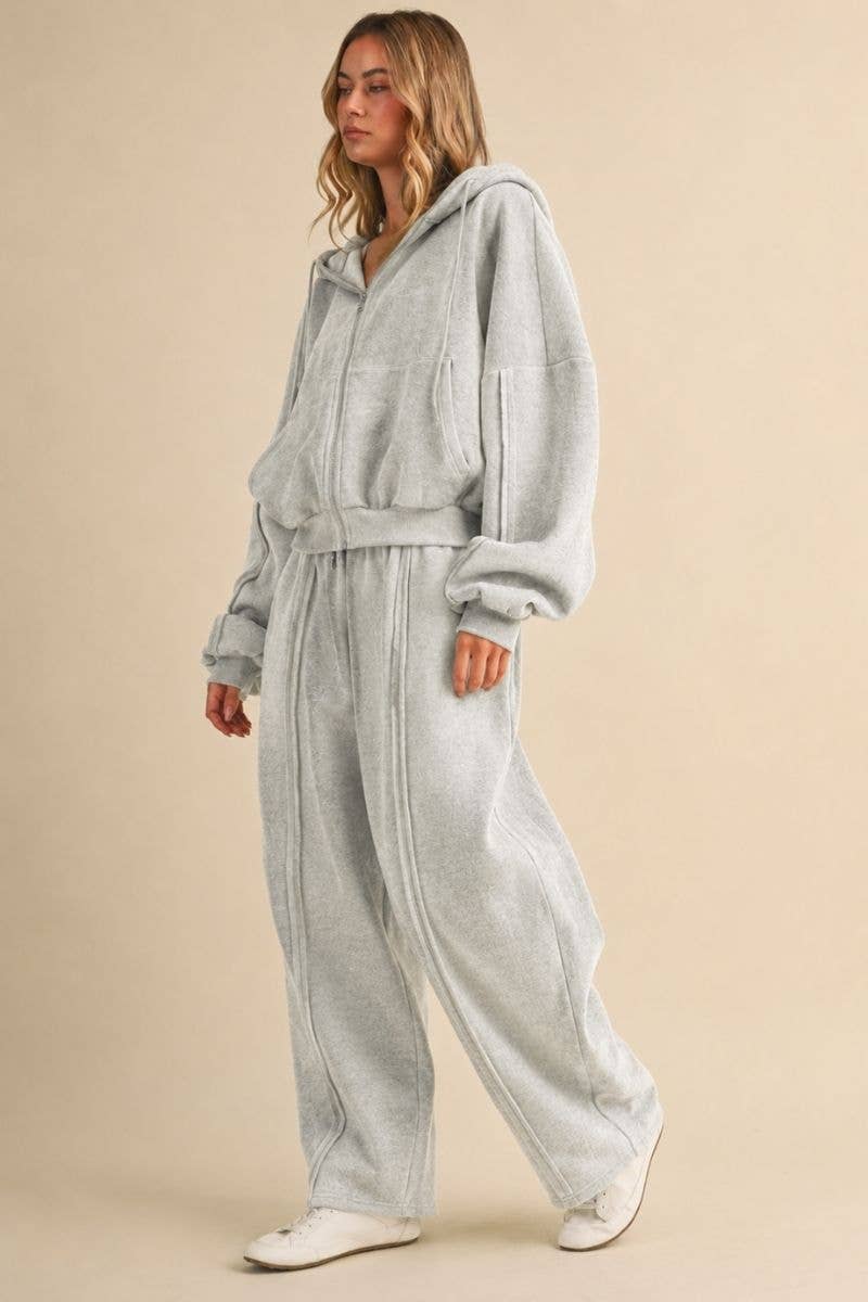 A woman modeling a light gray heathered lounge set consisting of a hooded sweatshirt and matching wide-leg sweatpants, photographed against a solid beige studio background.