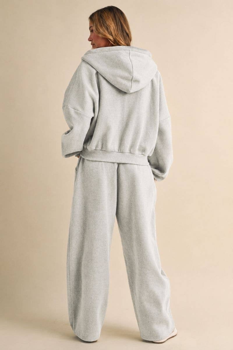 A woman wearing a light gray heathered hooded sweatshirt and matching wide-leg sweatpants, shown from the back against a plain beige studio background.