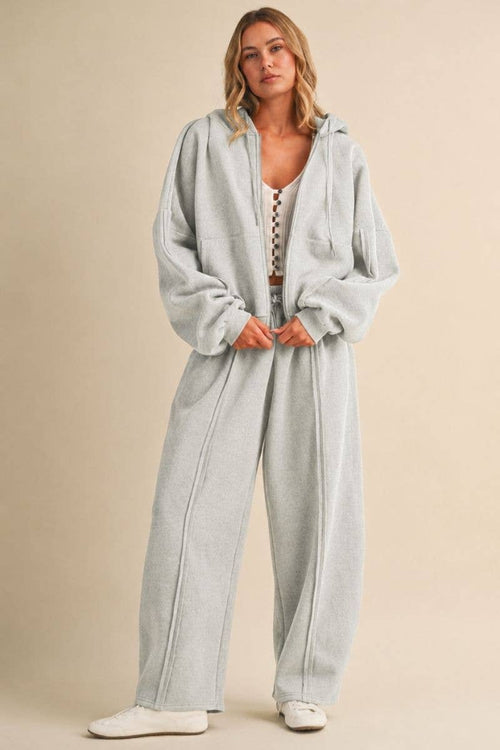 A woman modeling a light gray heathered hooded sweatshirt and matching wide-leg sweatpants set against a solid beige studio background.