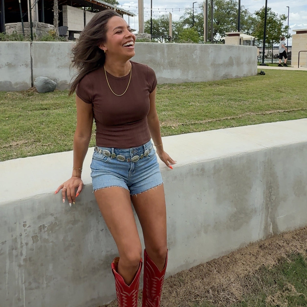 Person wearing red boots and denim shorts standing on a sidewalk with a blue sky and clouds in the background.