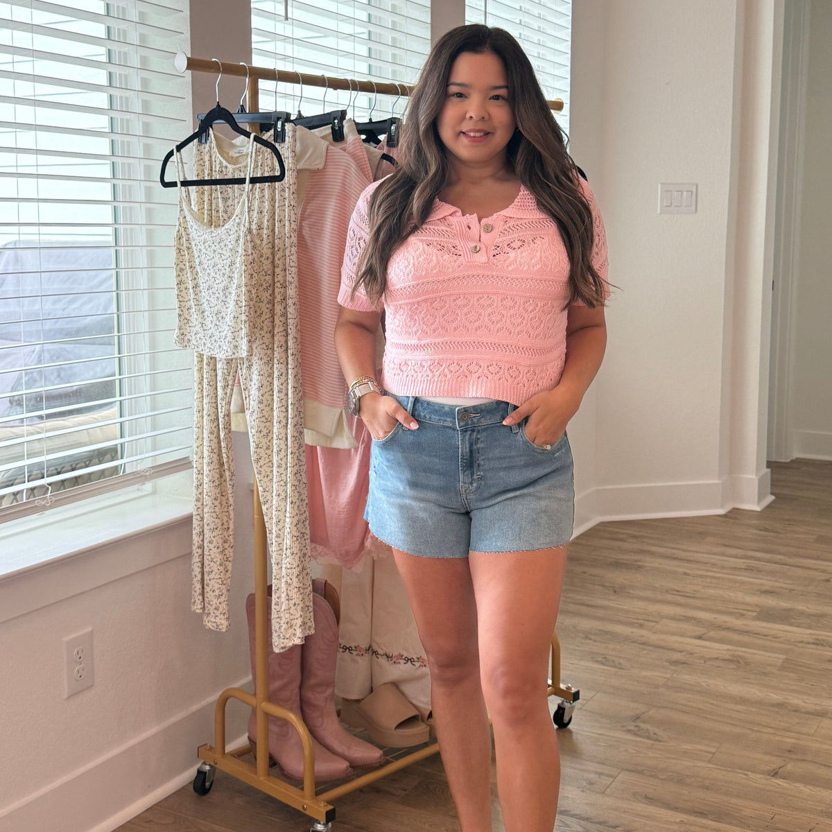 Woman standing in a room with a mirror reflecting her pink top and denim shorts.