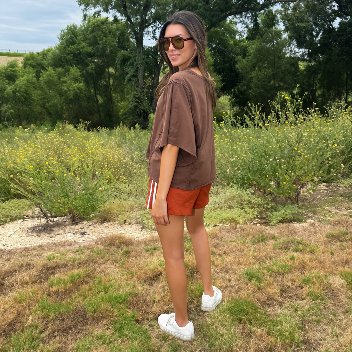 A woman wearing a brown oversized t-shirt, rust-orange shorts, white sneakers, fall fashion style , and stands outdoors in a grassy field with trees in the background.