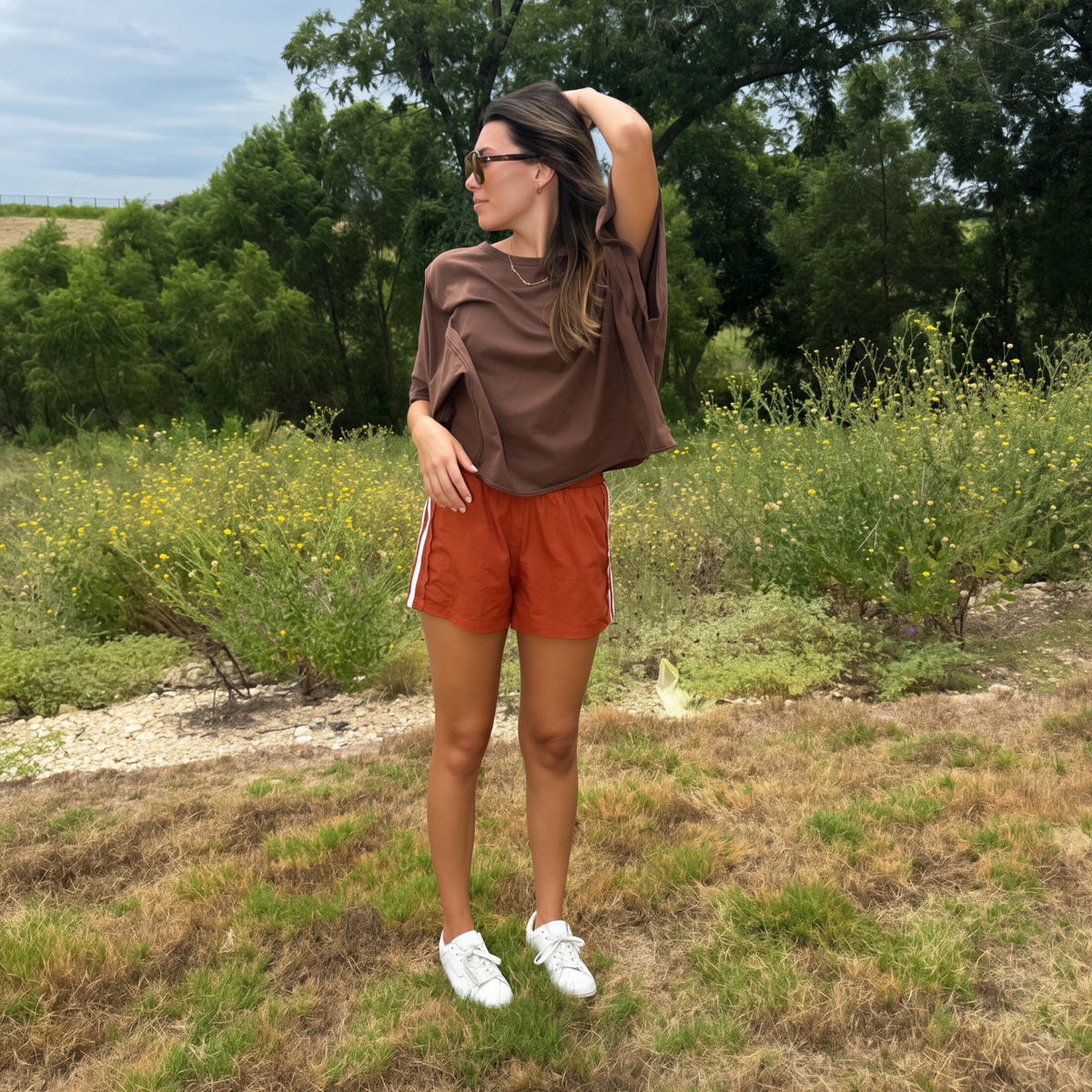 A woman stands outdoors in a grassy field with trees and shrubs in the background, wearing a loose brown t-shirt, rust-orange athletic shorts, and white sneakers.