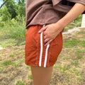 A person outdoors wearing Texas Burnt orange shorts with two white stripes down the side, paired with a brown top. The background shows grass and trees.