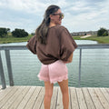 A woman stands on a wooden dock by a lake, wearing an oversized brown short-sleeve top, light pink shorts with lace trim, and white sneakers.
