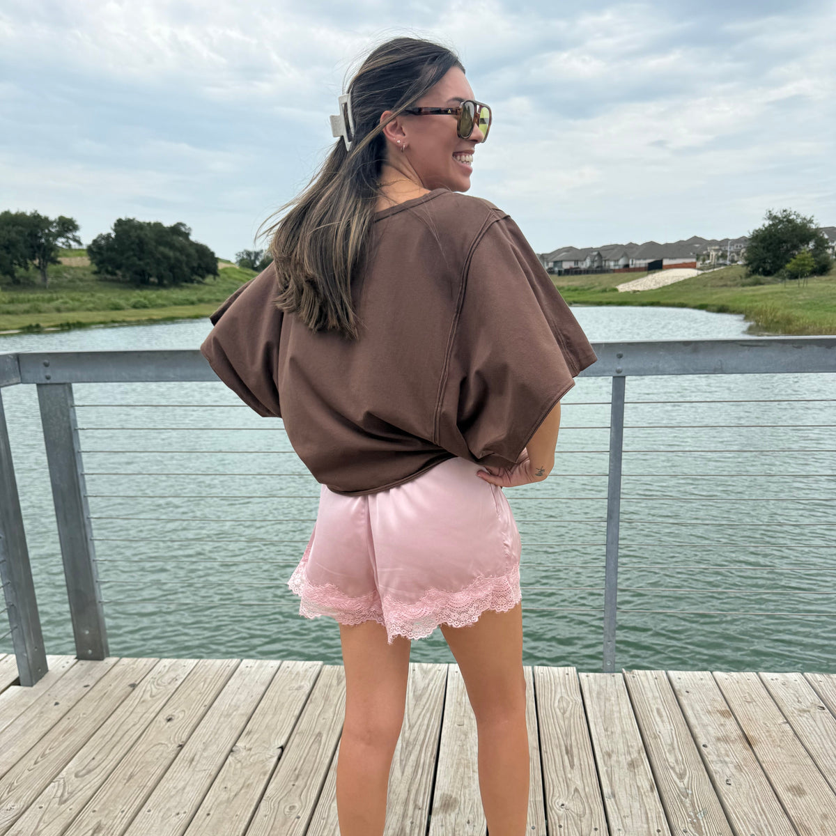 A woman stands on a wooden dock by a lake, wearing an oversized brown short-sleeve top, light pink shorts with lace trim, and white sneakers.