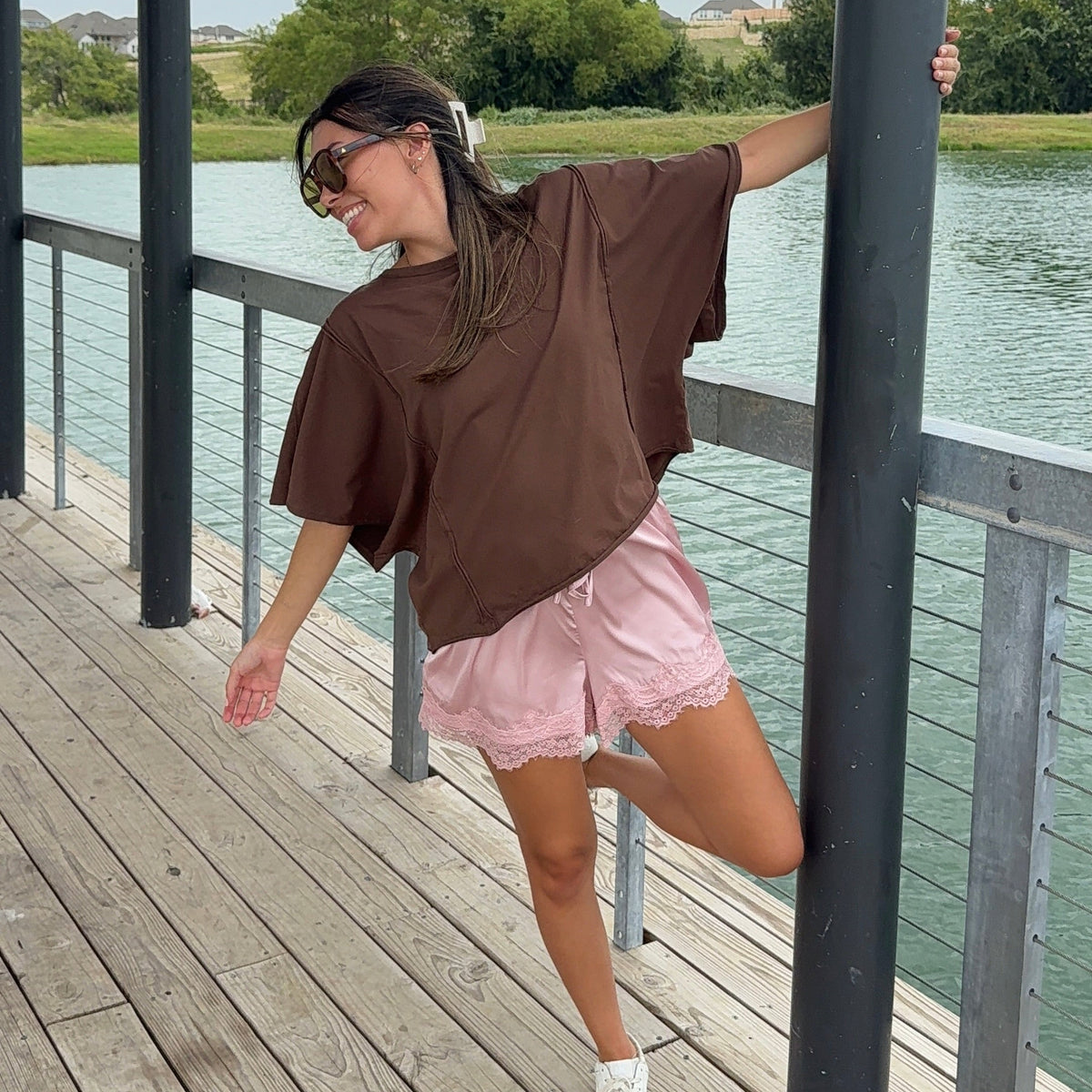 A woman posing on a wooden dock by a lake, wearing an oversized brown t-shirt, light pink shorts, and white sneakers.