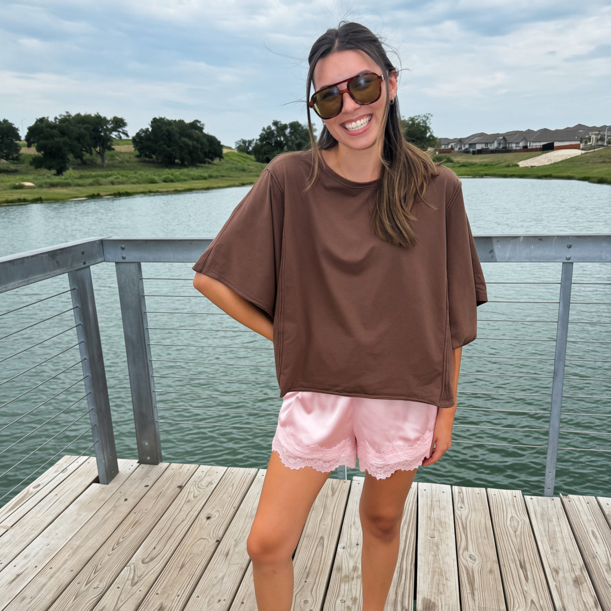 A woman stands on a wooden dock by a lake wearing an oversized brown t-shirt, light pink shorts, white sneakers, and sunglasses.