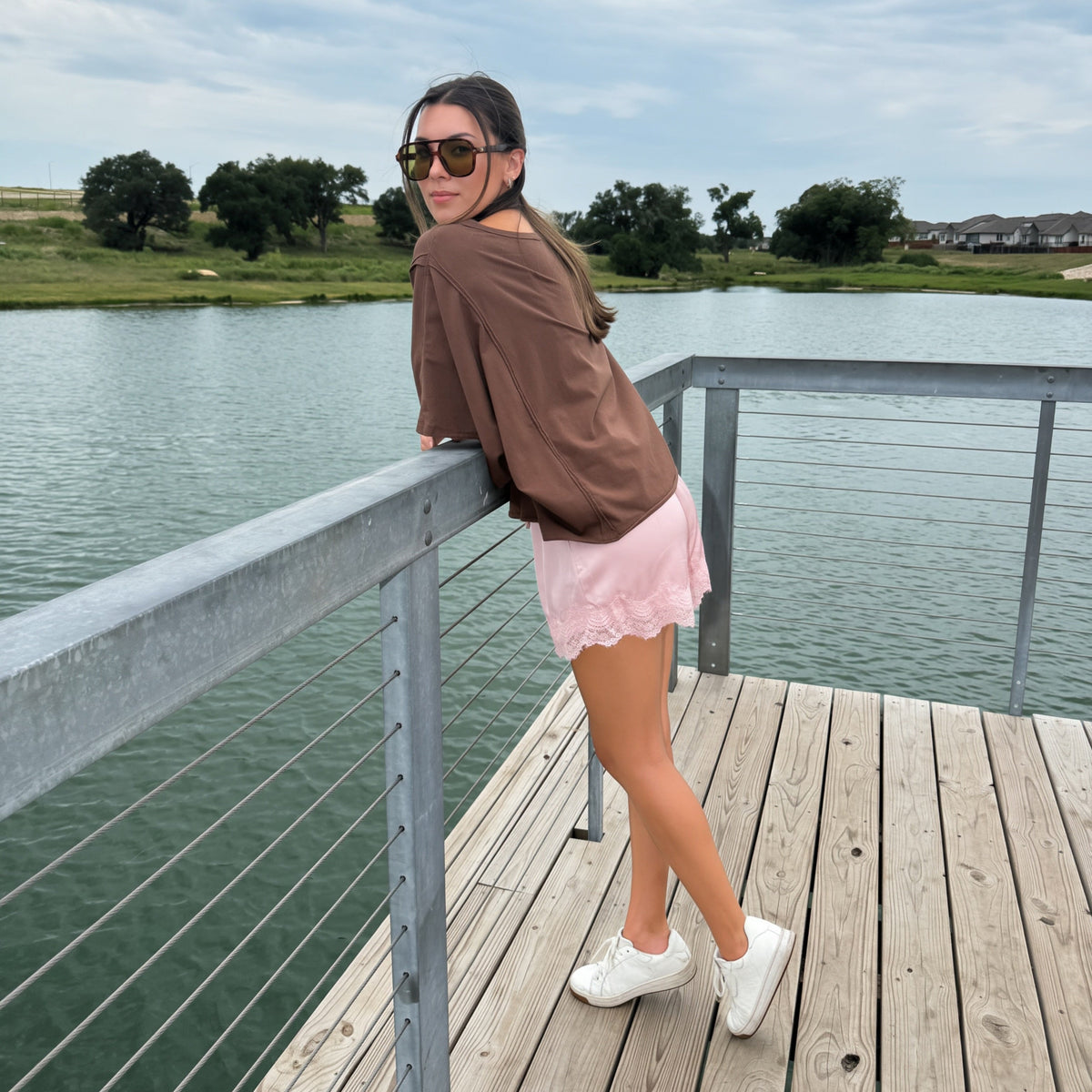 A woman wearing a brown off-the-shoulder top, a pink ruffled mini skirt, white sneakers, and sunglasses stands on a wooden dock by a lake, looking back over her shoulder.