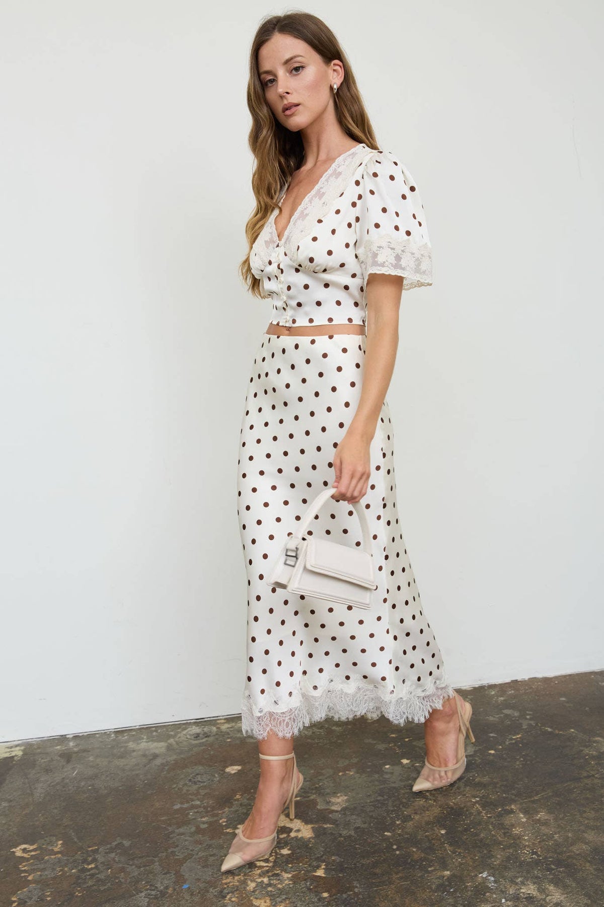 A woman models a white two-piece set featuring a short-sleeve wrap top and a matching midi skirt, both decorated with black polka dots and finished with white lace trim. She is holding a small white handbag and wearing beige high-heeled sandals against a white wall and concrete floor.