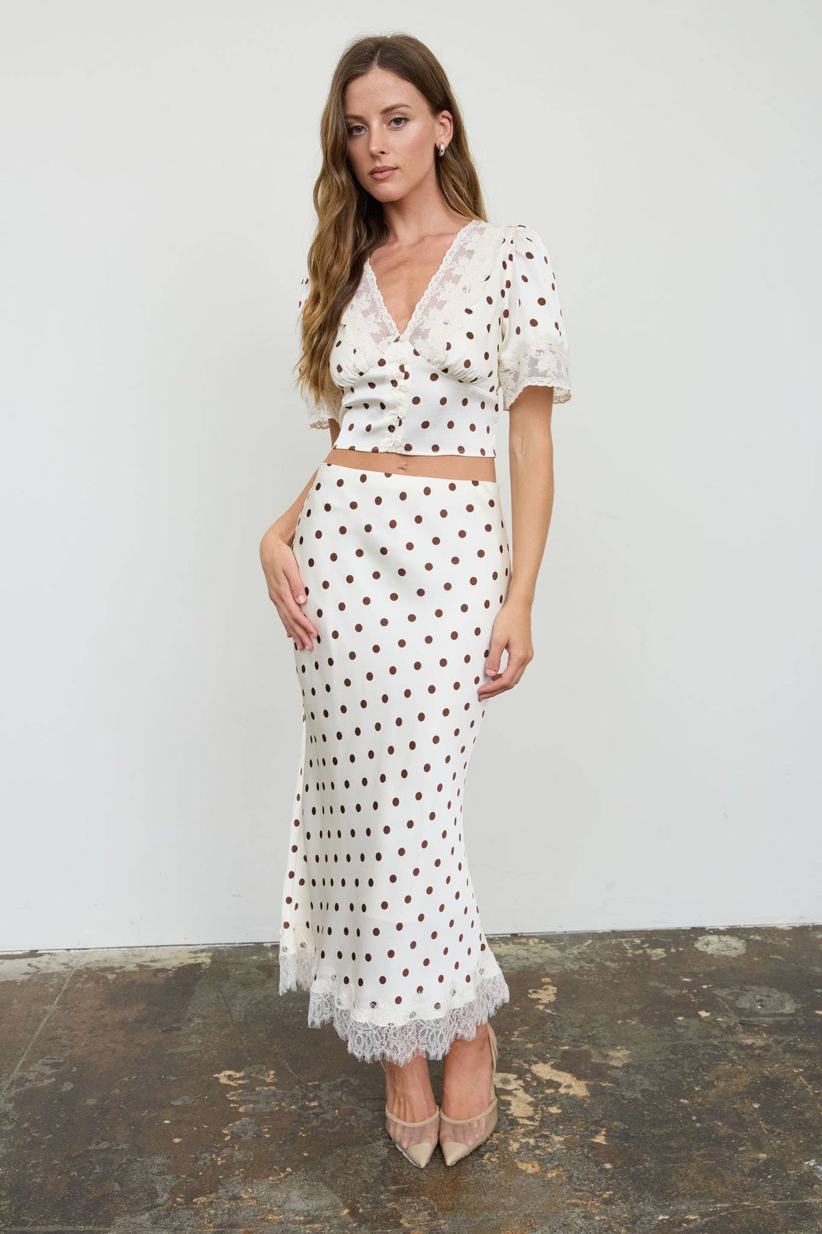 A woman models a two-piece white set with brown polka dots, featuring a short-sleeve wrap-style top with lace trim and a matching high-waisted maxi skirt with lace ruffles, set against a plain white wall.