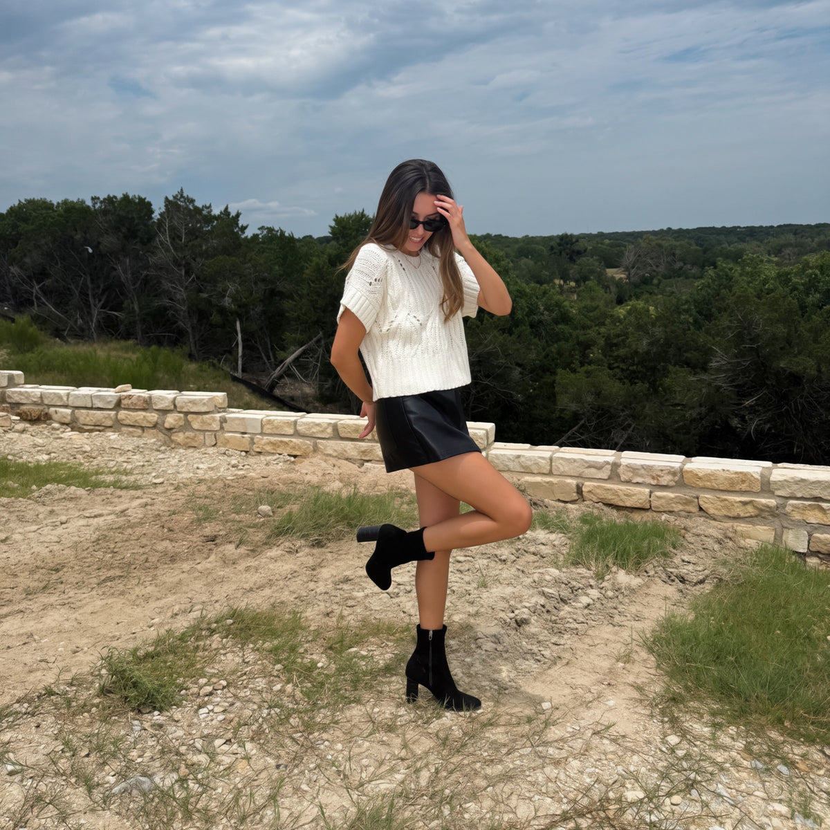 A woman posing outdoors on a dirt path with a stone wall and trees in the background, wearing a white textured short-sleeve top, a black leather mini skort, and black ankle boots.