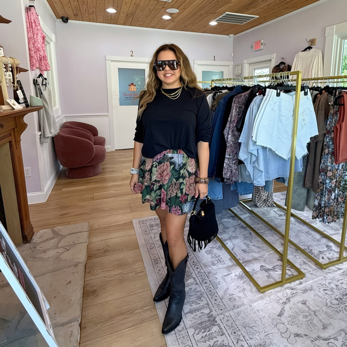 A woman posing in a boutique clothing store, wearing a black long-sleeve top, a floral print mini skort, and black knee-high boots. Clothing racks and a crystal chandelier are visible in the background.