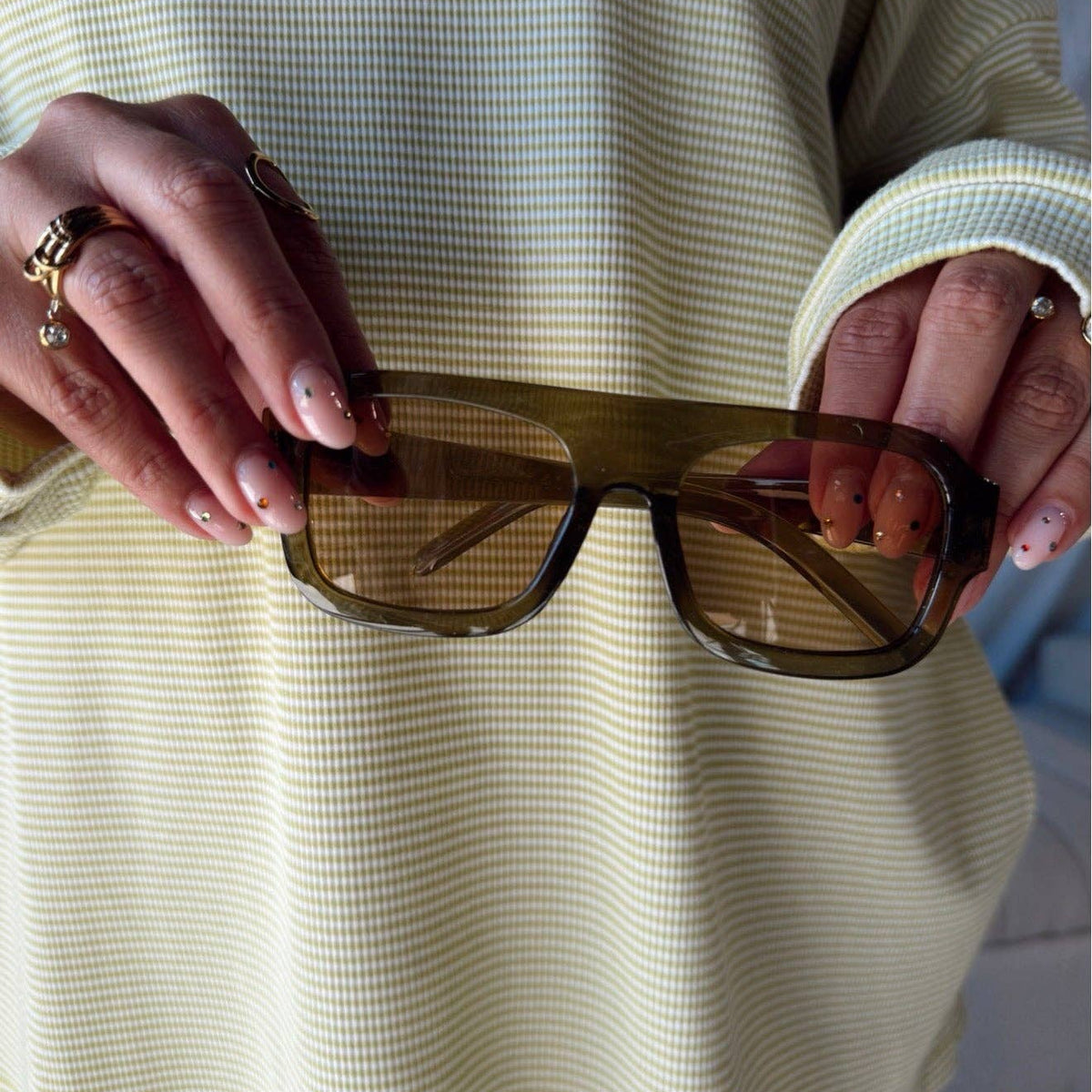 A person holding a pair of translucent dark brown square sunglasses in front of a light green and white gingham shirt.