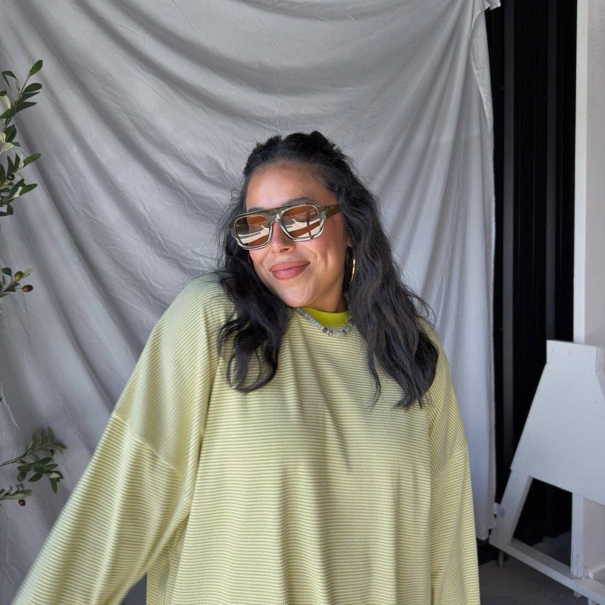 A woman wearing a pale yellow-green oversized ribbed long-sleeve top and chunky square sunglasses, standing in front of a draped white fabric backdrop.