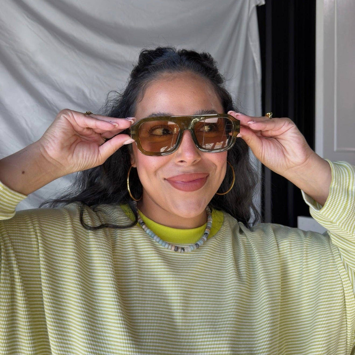 A woman wearing oversized square sunglasses with a translucent amber-brown frame and tinted lenses, holding them up to her eyes. She is wearing a pale yellow-green ribbed long-sleeve top, gold hoop earrings, and a beaded necklace, with a draped white fabric backdrop behind her.