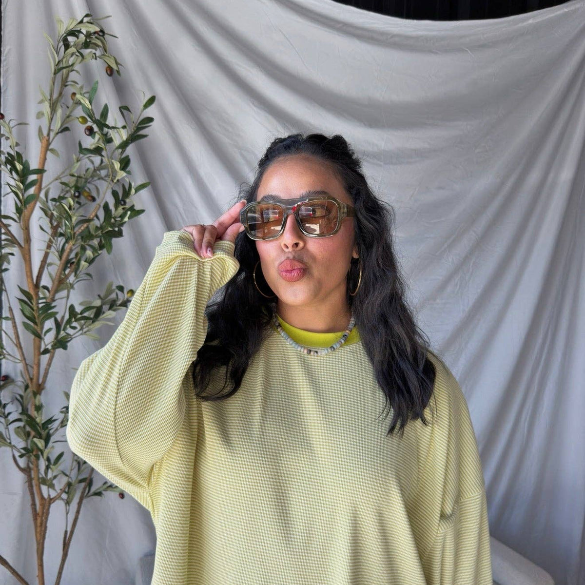A woman wearing a pale yellow ribbed knit long-sleeve top and oversized tortoiseshell sunglasses, posing in front of a draped white fabric backdrop with a potted olive tree on the left.
