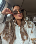 A woman wearing oversized square sunglasses with a tortoiseshell pattern and a white sherpa fleece jacket with brown graphic details, sitting inside a car.