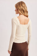 A woman shown from the back wearing a fitted cream long-sleeve top with lace trim around the neckline and a brown satin skirt, against a plain light gray studio background.
