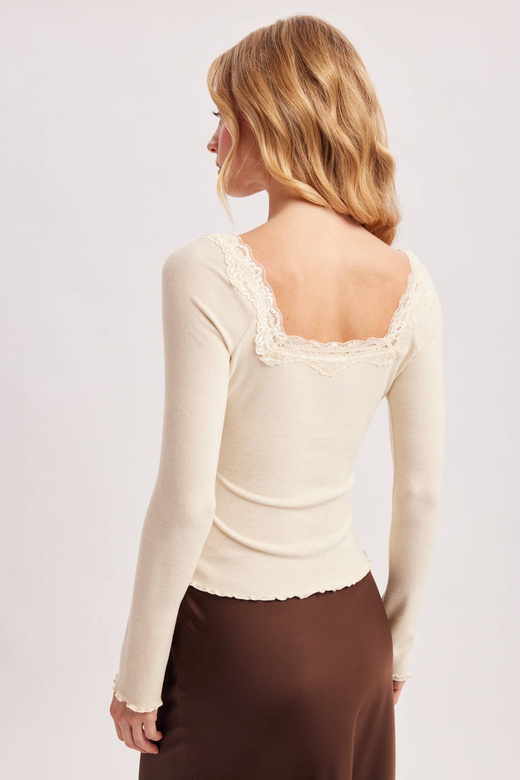 A woman shown from the back wearing a fitted cream long-sleeve top with lace trim around the neckline and a brown satin skirt, against a plain light gray studio background.