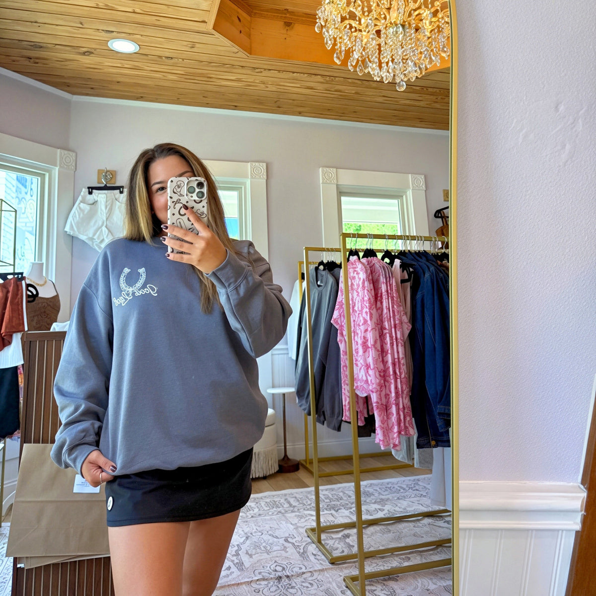 A woman takes a mirror selfie in a boutique, wearing a slate blue-gray sweatshirt with a white embroidered graphic and black shorts. The boutique features a wood-paneled ceiling, a crystal chandelier reflection, and clothing racks in the background.
