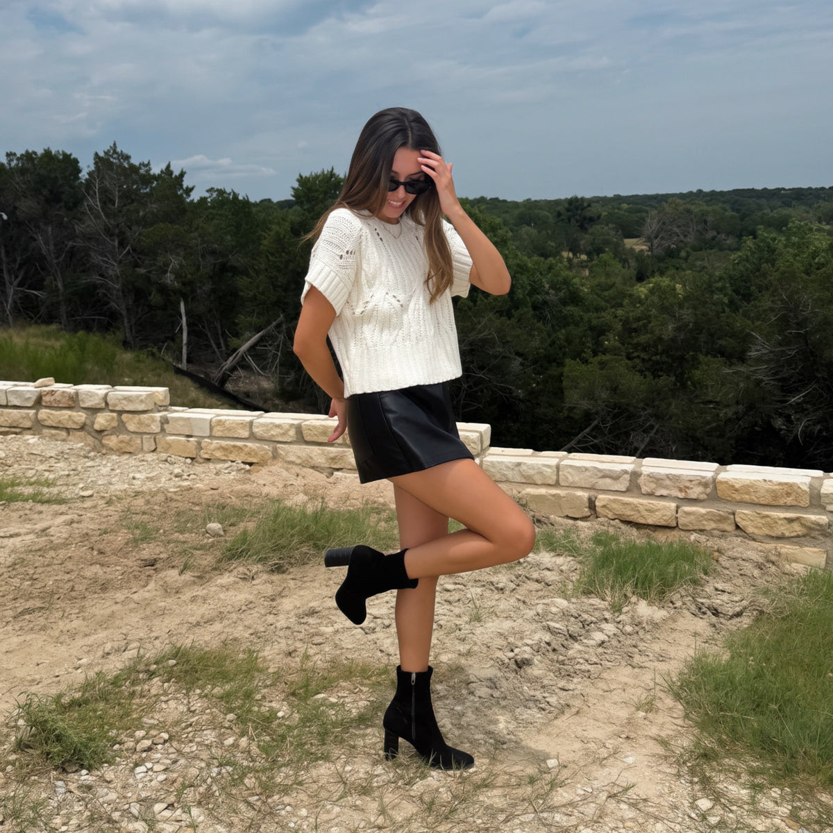 A woman posing outdoors on a dirt path with a stone wall and trees in the background, wearing a white textured short-sleeve top, a black leather mini skirt, and black ankle boots.