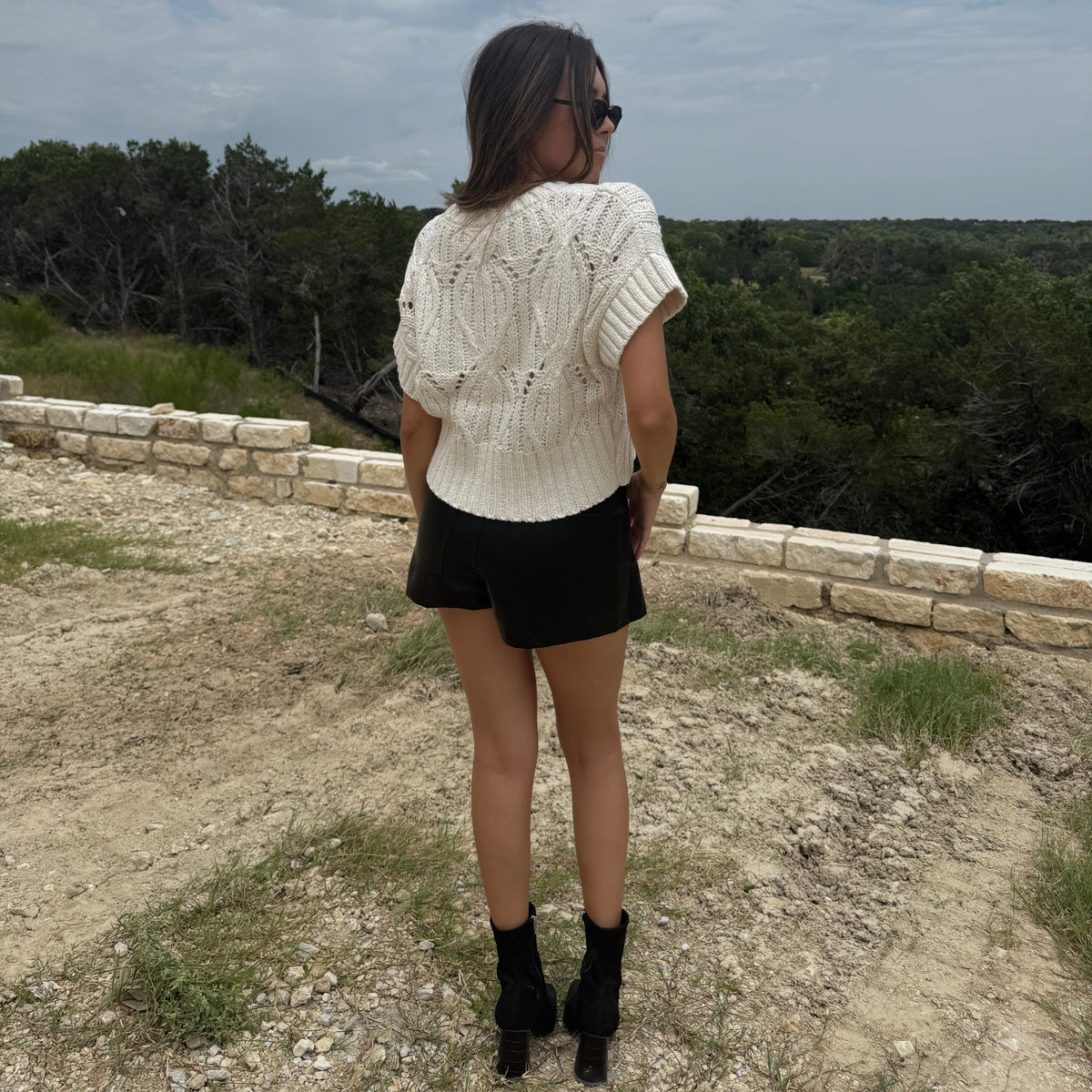 A woman is seen from behind wearing a cream-colored textured knit short-sleeve top, black shorts, and black ankle boots, standing outdoors on a dirt path with a stone wall and trees in the background under a cloudy sky.