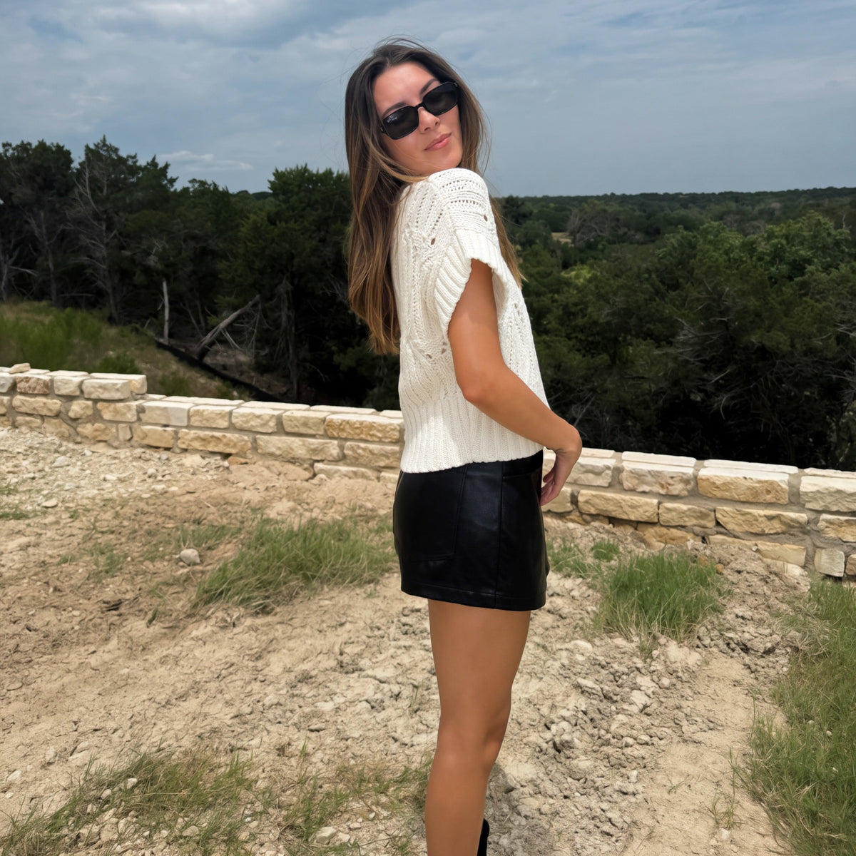 A woman wearing a white textured short-sleeve top and a black faux-leather mini skirt, standing outdoors on a dirt path with a stone wall and trees in the background.