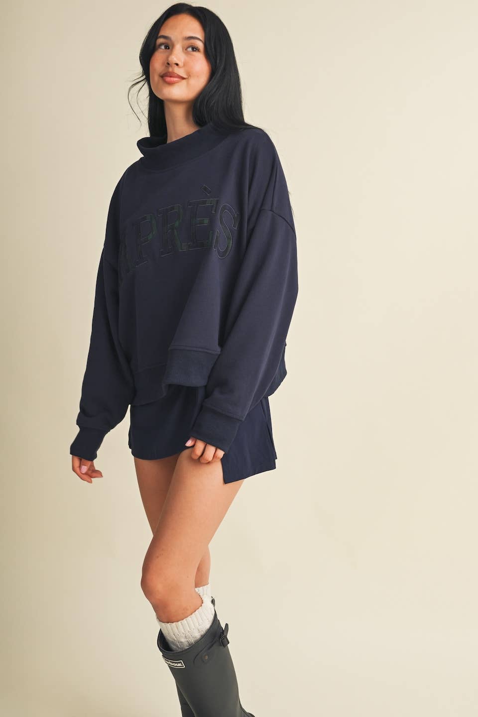 A woman modeling an oversized navy blue sweatshirt with a tonal 'APRÈS' logo across the chest, paired with white socks and dark rain boots, against a solid light beige background.