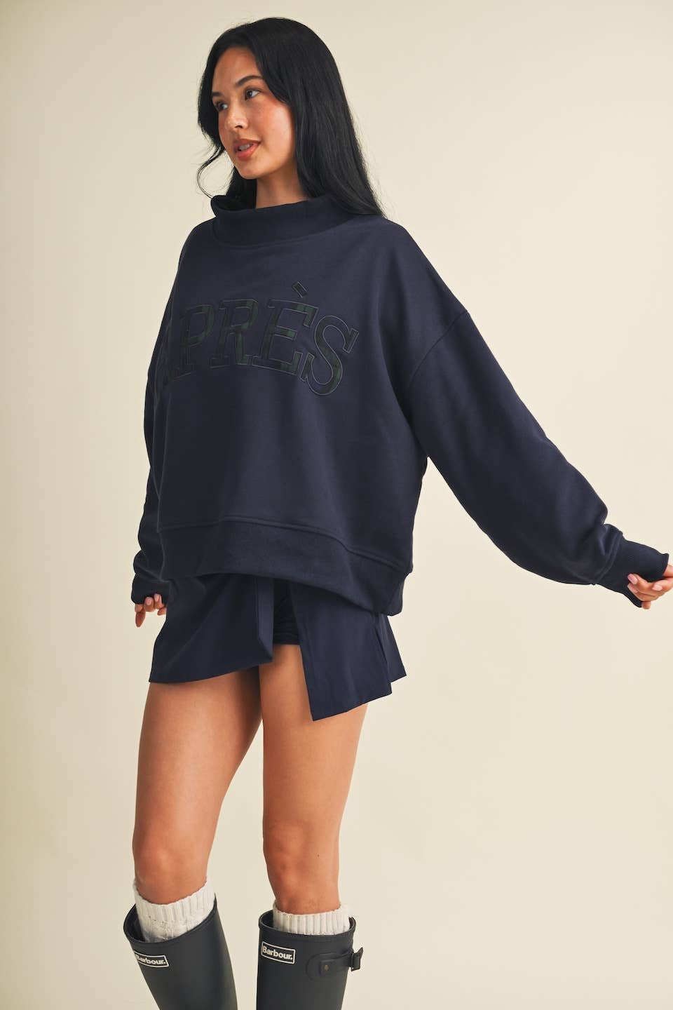 A woman modeling an oversized navy blue sweatshirt with a tonal 'PRESS' logo across the chest, paired with matching navy shorts and black rain boots, against a solid beige background.