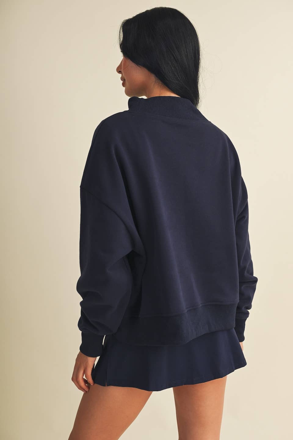 A woman shown from the back wearing an oversized navy blue sweatshirt and matching shorts against a plain beige studio background.