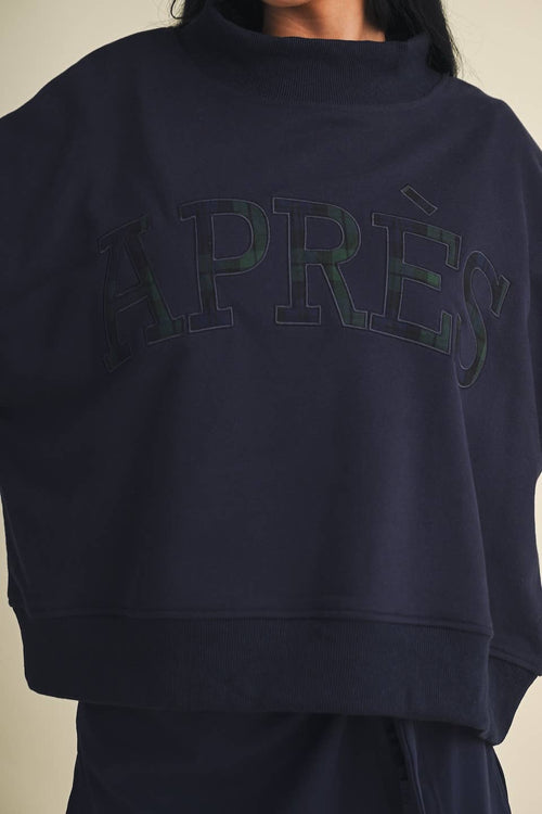 A close-up of a person wearing a dark navy crewneck sweatshirt with 'APRÈS' embroidered across the chest in a tonal green and black pattern.