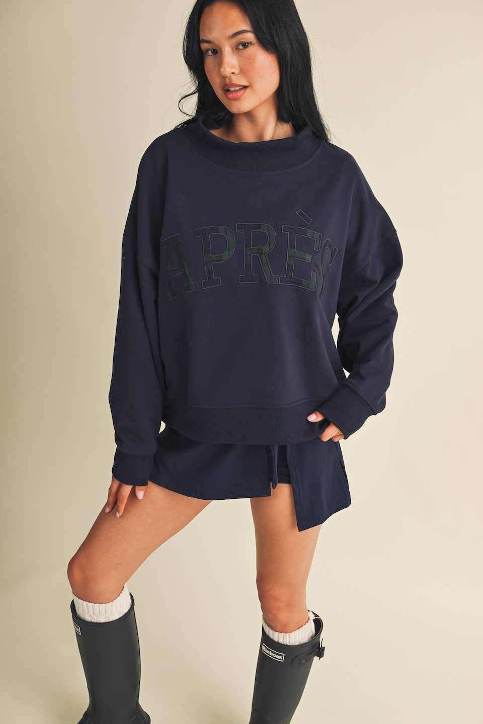 A woman modeling an oversized navy blue sweatshirt with a tonal dark logo across the chest, paired with a matching navy mini skirt and black knee-high boots against a plain beige background.