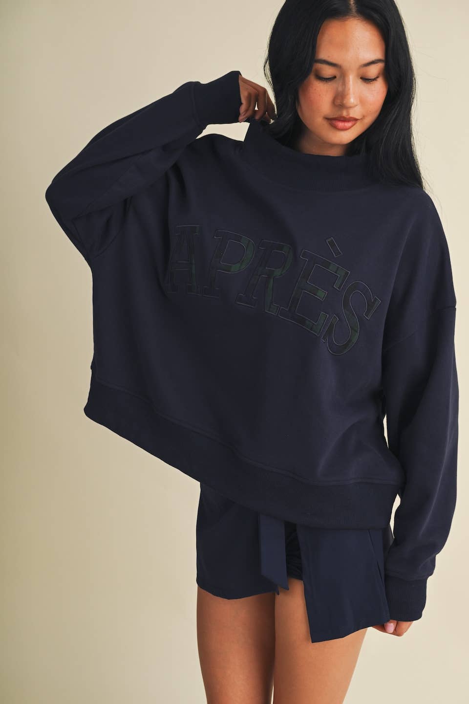 A woman wearing a dark navy oversized sweatshirt with tonal 'APRÈS' lettering across the chest, paired with matching shorts, against a plain beige studio background.