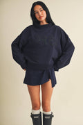 A woman modeling an oversized navy blue sweatshirt with a matching mini skirt, styled with white socks and black rain boots against a plain beige background.