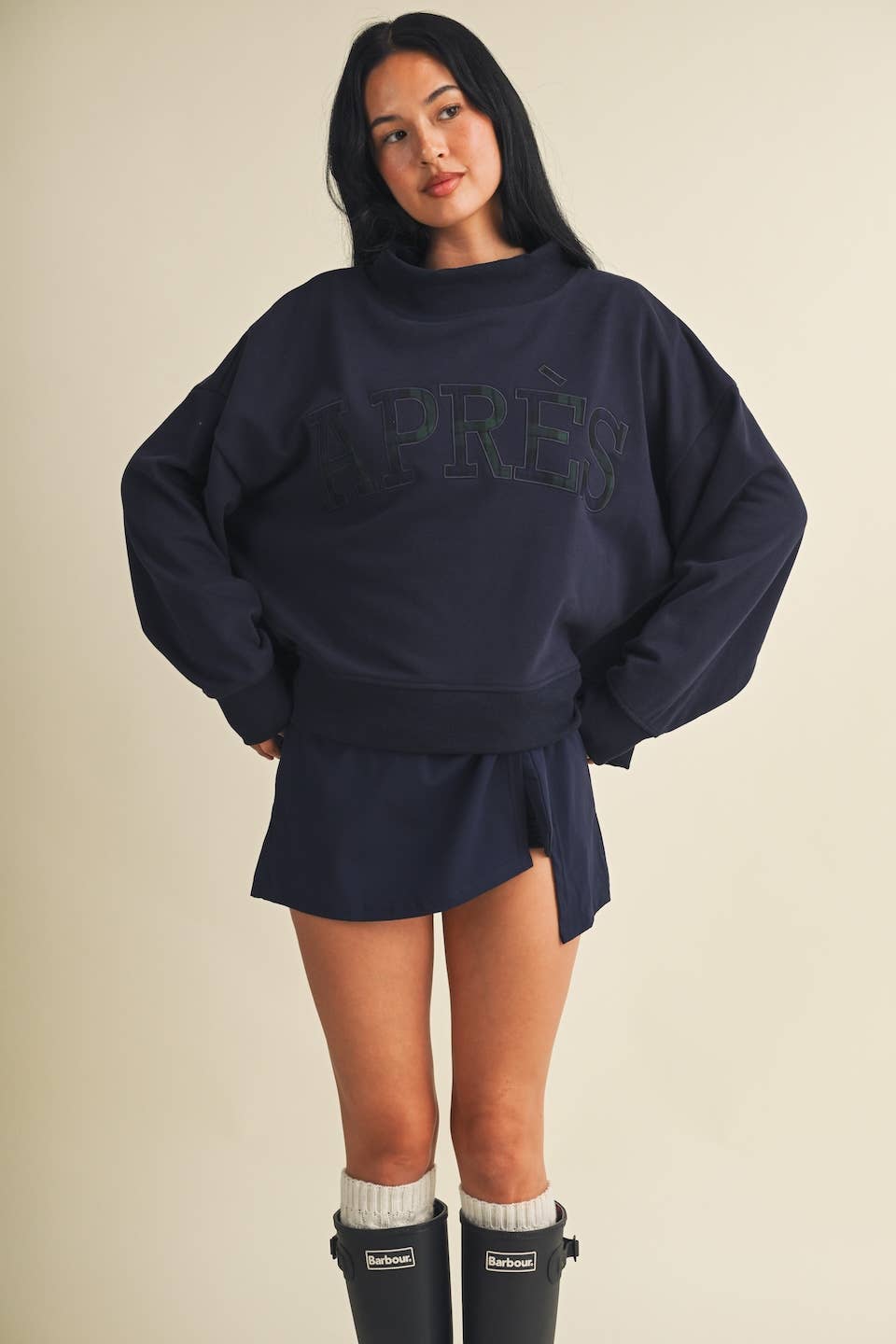 A woman modeling an oversized navy blue sweatshirt with a matching mini skirt, styled with white socks and black rain boots against a plain beige background.