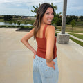 A woman wearing a burnt orange colored ribbed tank top and light blue jeans with gold star details, standing outdoors on a concrete patio with a lake and trees in the background.