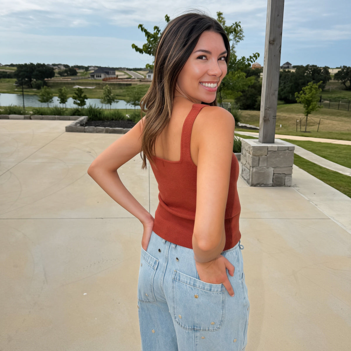 A woman wearing a burnt orange colored ribbed tank top and light blue jeans with gold star details, standing outdoors on a concrete patio with a lake and trees in the background.