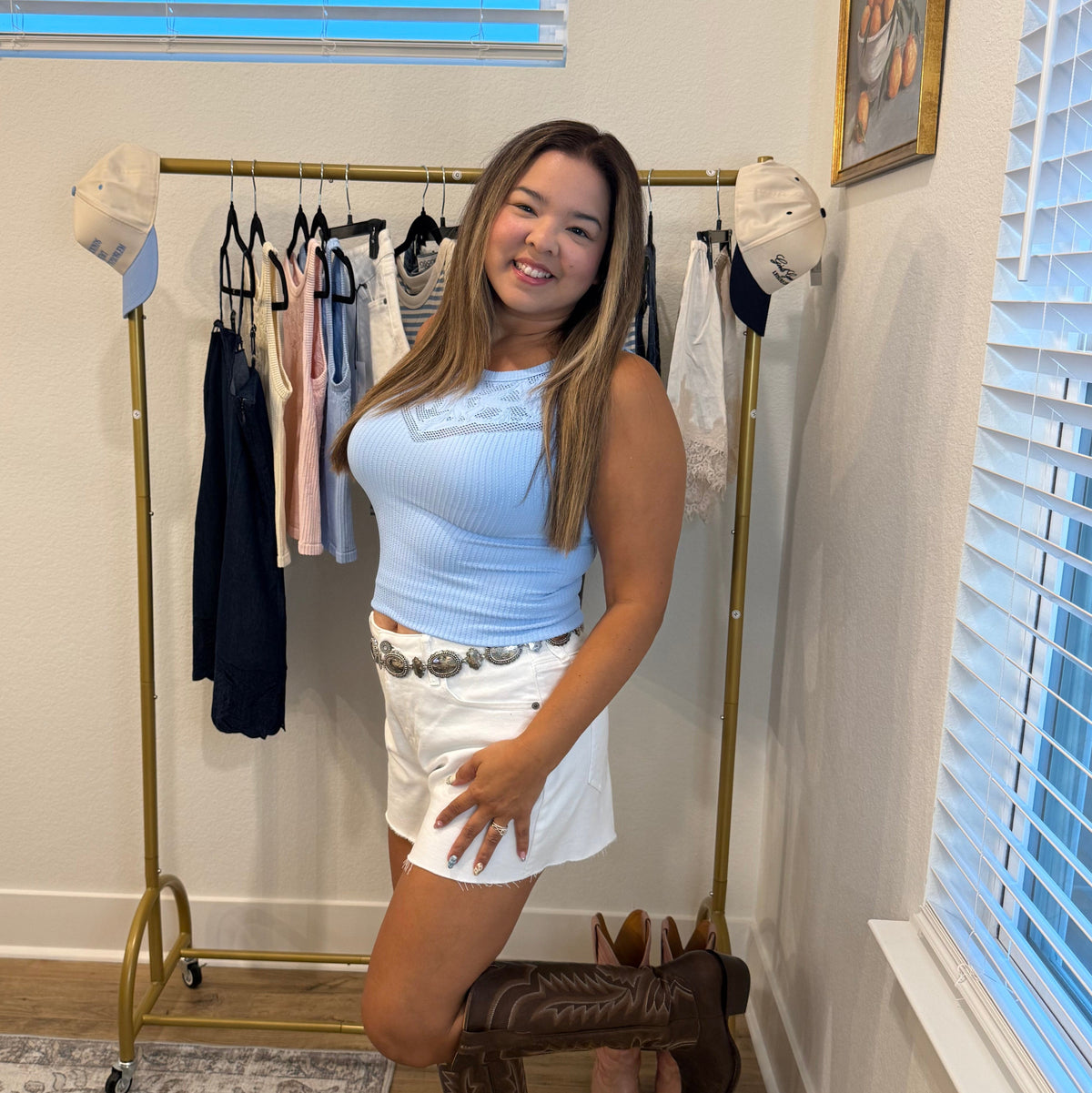 A woman posing in a white sleeveless top and white shorts with a silver belt, paired with brown cowboy boots, standing in a room with a clothing rack and window blinds.