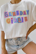 A person wearing a white graphic T-shirt with colorful patchwork-style letters spelling 'GAMEDAY GIRLIE' and light-wash denim shorts against a plain light background.