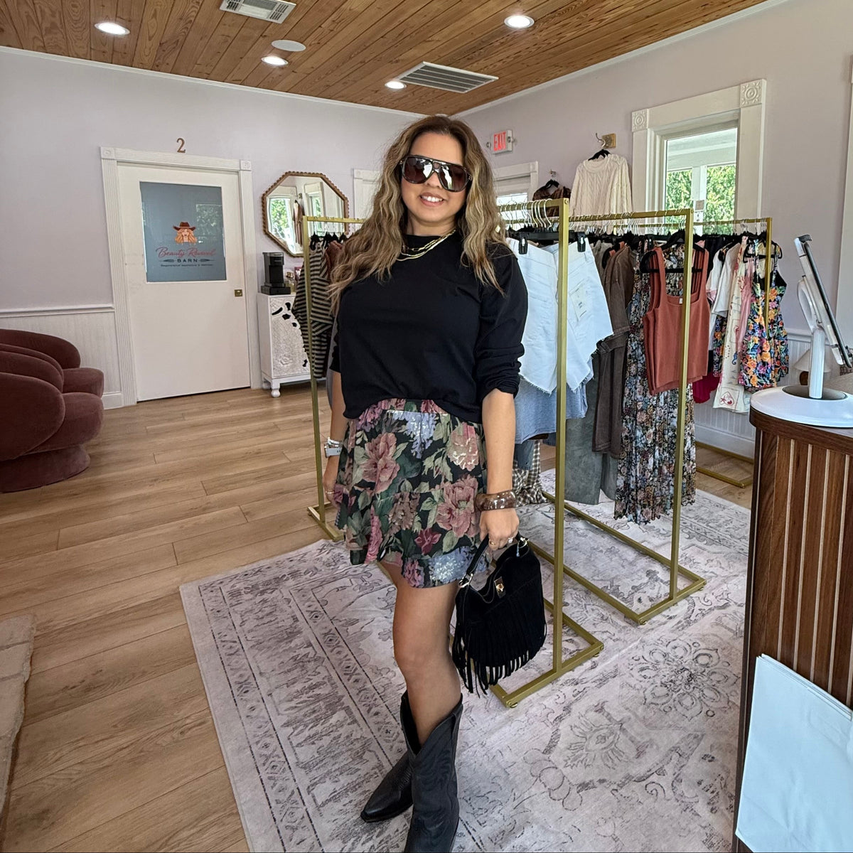 A woman posing in a boutique shop wearing a black long-sleeve top, a colorful floral mini skirt, black cowboy boots, and sunglasses. Clothing racks, a wooden ceiling, and a chandelier are visible in the background.