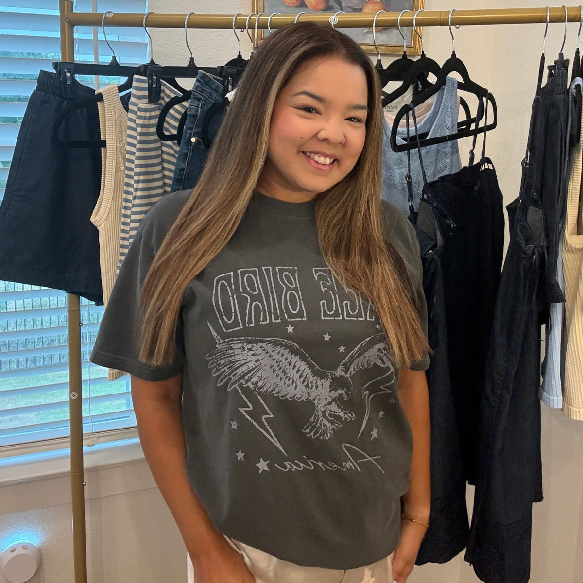 A smiling young woman stands indoors in front of a clothing rack, wearing a dark gray graphic T-shirt with an eagle design and the words 'BIRD' and 'America' visible on the front, paired with light beige pants.