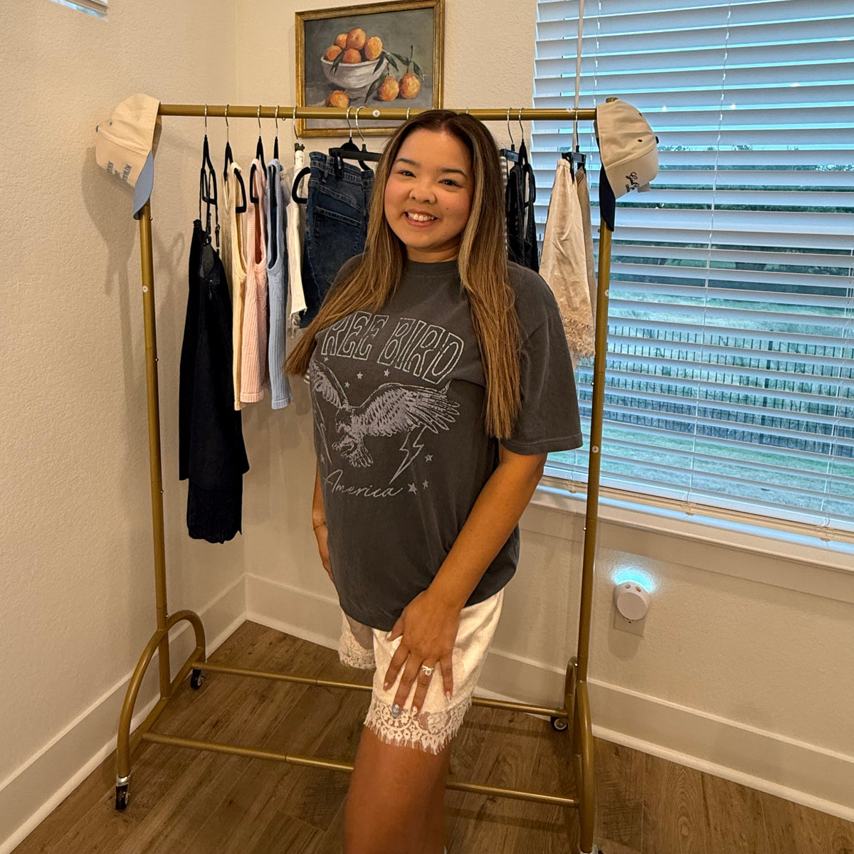 A woman stands in a room wearing a dark gray graphic t-shirt and white lace-trimmed shorts, posing next to a gold clothing rack filled with various garments.